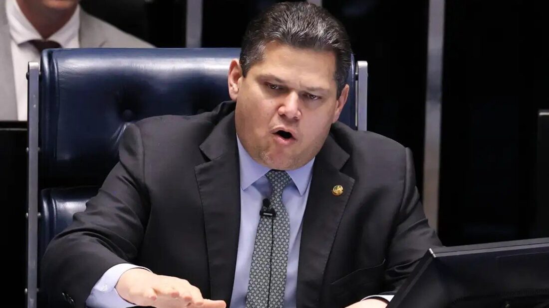 O senador Davi Alcolumbre, de terno e gravata, sentado na cadeira da presidência do Senado, fala com a mão estendida sobre a mesa