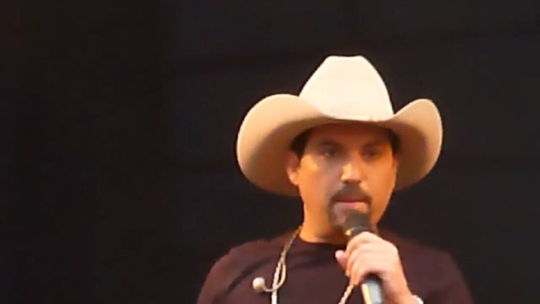 Cantor de chapéu claro e camiseta preta canta ao microfone em palco com fundo escuro