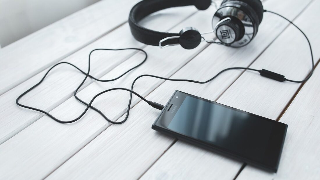 Fone de ouvido preto conectado por fio a um celular sobre uma mesa de madeira branca