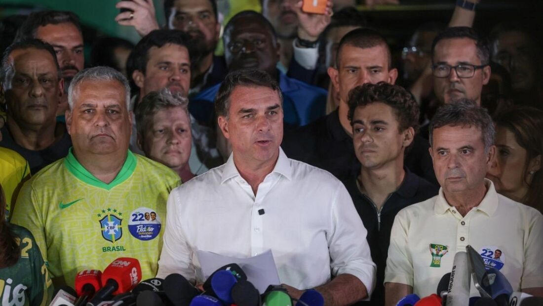 O senador Flávio Bolsonaro, de camisa branca, fala a jornalistas cercado de apoiadores, alguns com camisas da seleção, diante de vários microfones