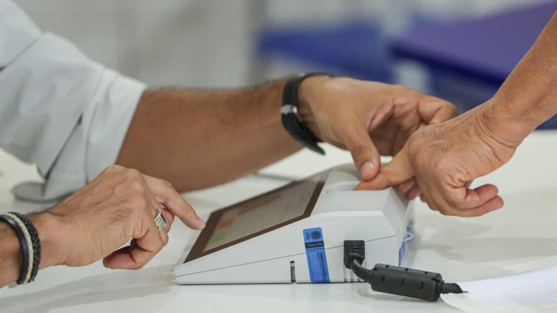 Eleitor coloca o dedo no leitor biométrico diante de mesário em seção eleitoral
