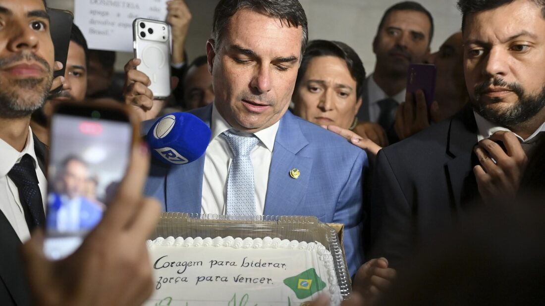 O senador Flávio Bolsonaro, de terno azul, cercado de pessoas e celulares, segura um bolo com a inscrição 'Coragem para liderar, força para vencer'