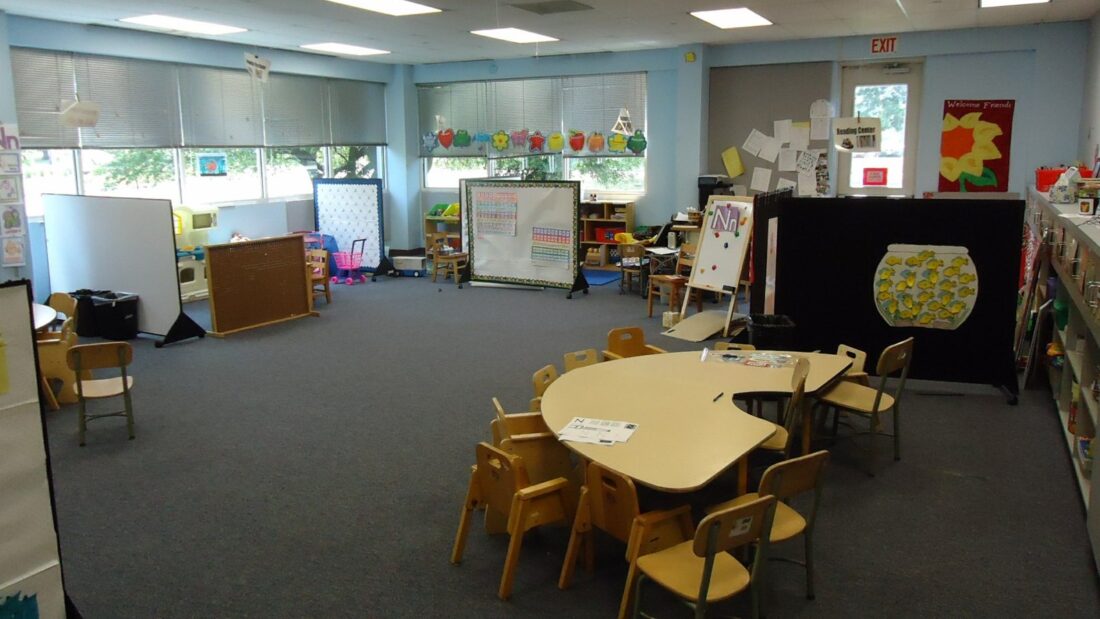 Sala de educação infantil vazia, com mesas baixas, cadeiras pequenas e brinquedos