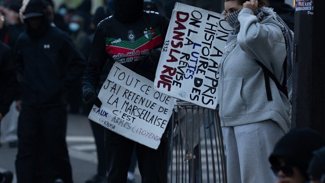 Manifestantes mascarados e de capuz seguram cartazes escritos à mão ao lado de uma lixeira em chamas numa calçada de Paris
