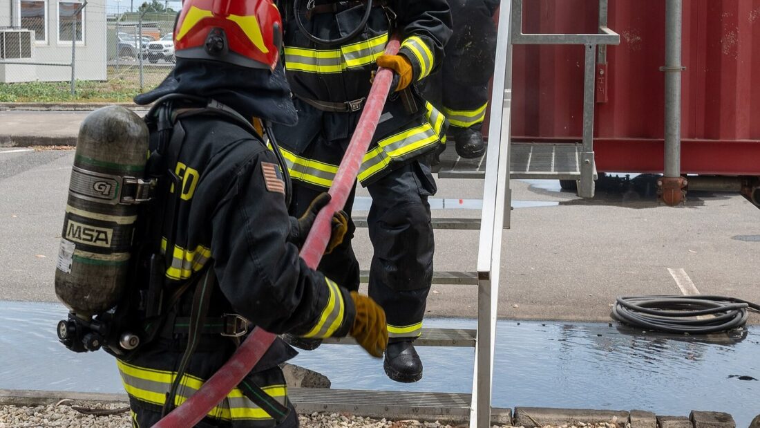 Dois bombeiros com roupa de proteção, capacete e cilindro de ar sobem a escada de um contêiner de treinamento segurando uma mangueira, com fumaça saindo da porta