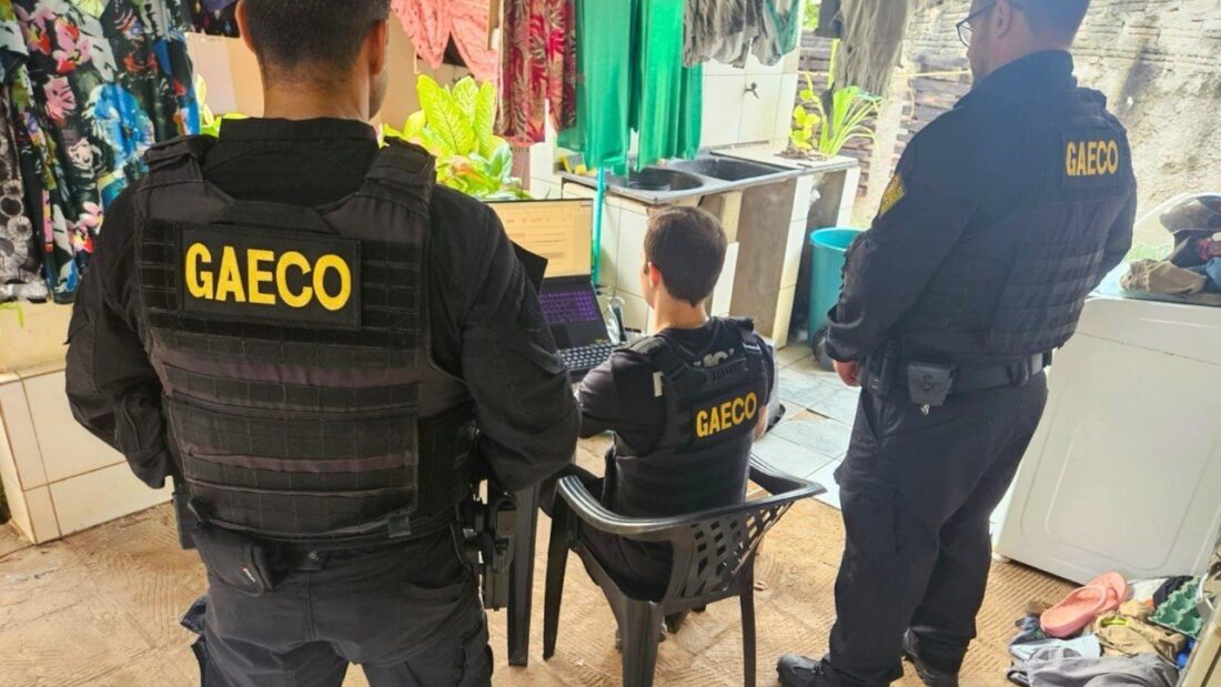 Três agentes do Gaeco, de colete preto com a palavra Gaeco nas costas, analisam material em um computador durante cumprimento de mandado dentro de um imóvel