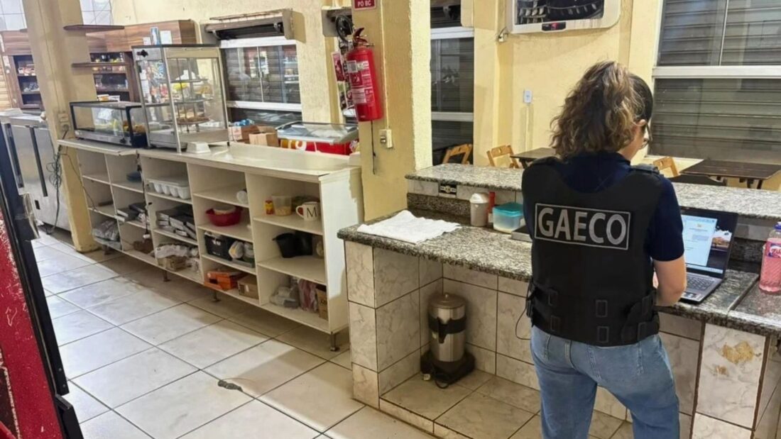 Agente com colete do Gaeco, de costas, examina um computador portátil sobre o balcão de uma cozinha industrial