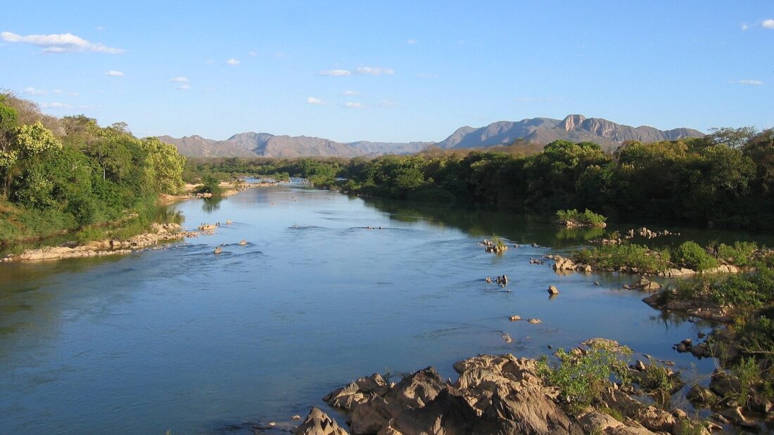 Rio Paranã corre entre pedras e margens de mata, com serras ao fundo, sob céu azul