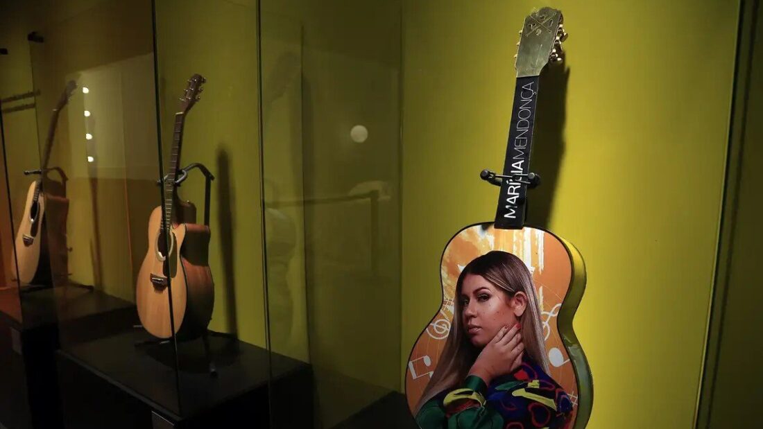 Violões expostos em vitrine iluminada; um deles tem o retrato de Marília Mendonça pintado no corpo e o nome dela no braço