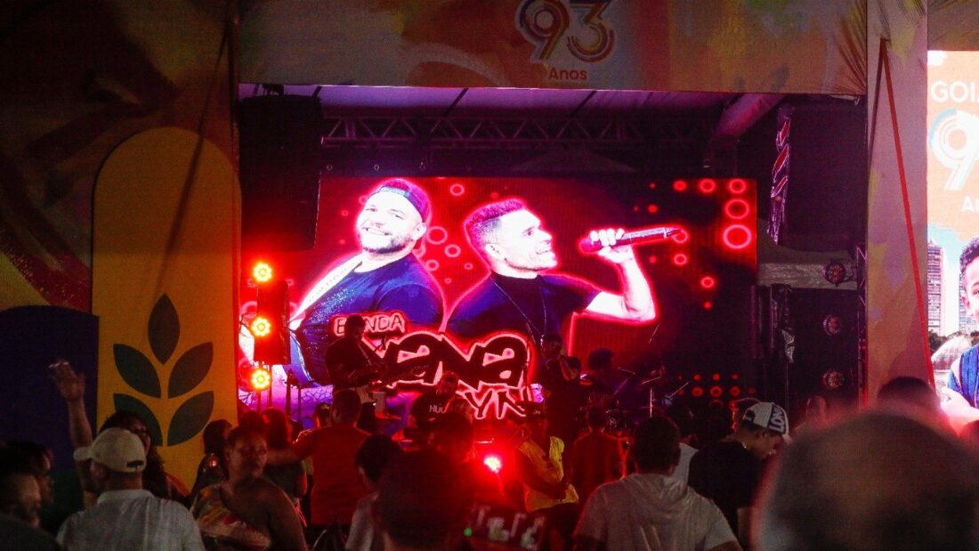 Palco iluminado de vermelho com telão mostrando cantores, decoração com o número 93 de Goiânia e público assistindo à noite
