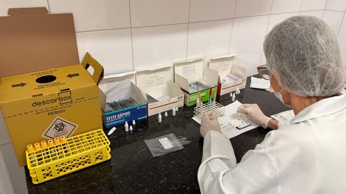 Profissional de saúde de jaleco branco, touca e máscara manipula testes rápidos numa bancada de laboratório, ao lado de uma caixa coletora de material perfurocortante e de um suporte amarelo com tubos