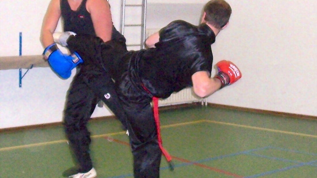 Dois lutadores treinam em uma quadra coberta; um deles, de uniforme preto e luvas vermelhas, aplica um chute lateral enquanto o outro, de luvas azuis, se defende