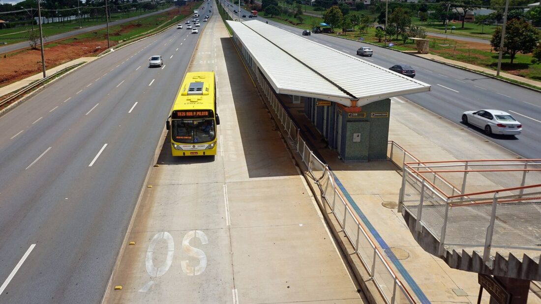 Ônibus amarelo do BRT em corredor exclusivo ao lado de uma estação coberta, entre as pistas de uma avenida larga em Brasília