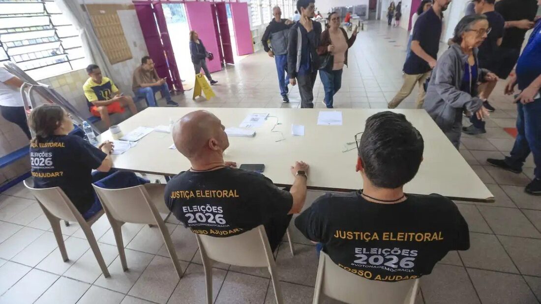Mesários com camisetas da Justiça Eleitoral, sentados a uma mesa no pátio de uma escola, enquanto eleitores circulam ao fundo