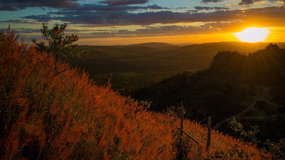 Pôr do sol visto do alto do Pico dos Pireneus, com o sol entre nuvens sobre serras escuras e, em primeiro plano, a vegetação do Cerrado iluminada em tons de laranja