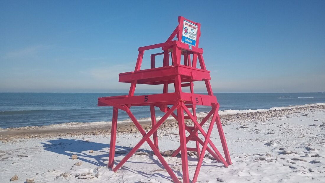 Cadeira alta de guarda-vidas pintada de vermelho, vazia, sobre a areia coberta de neve, com a água do lago e o céu azul ao fundo