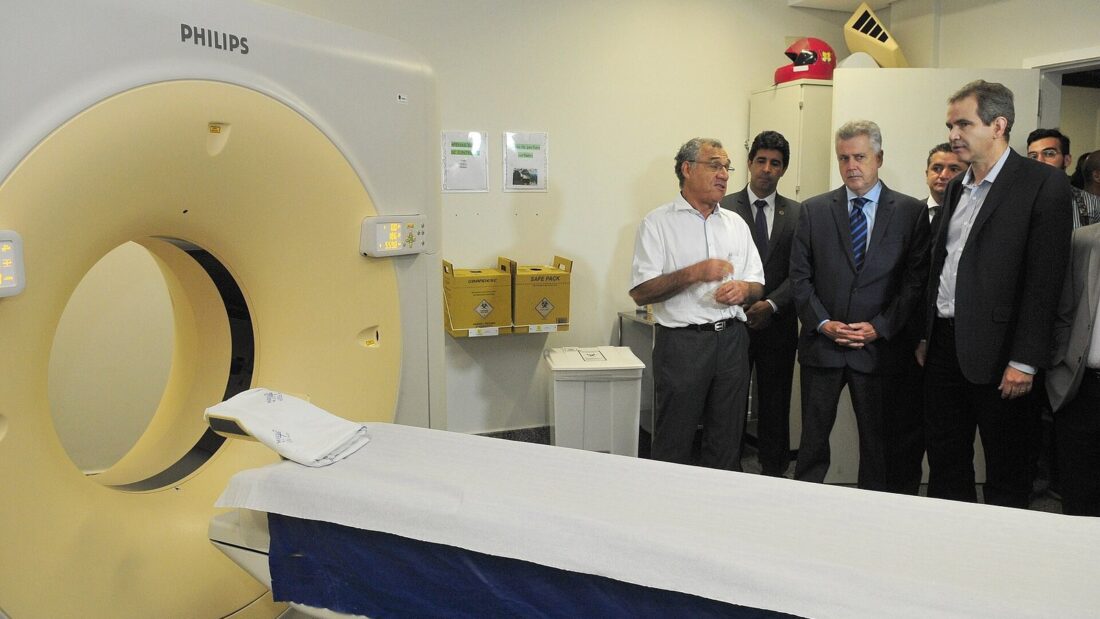 Sala de tomografia do Hugol, com o equipamento em primeiro plano e um grupo de homens de terno conversando ao fundo