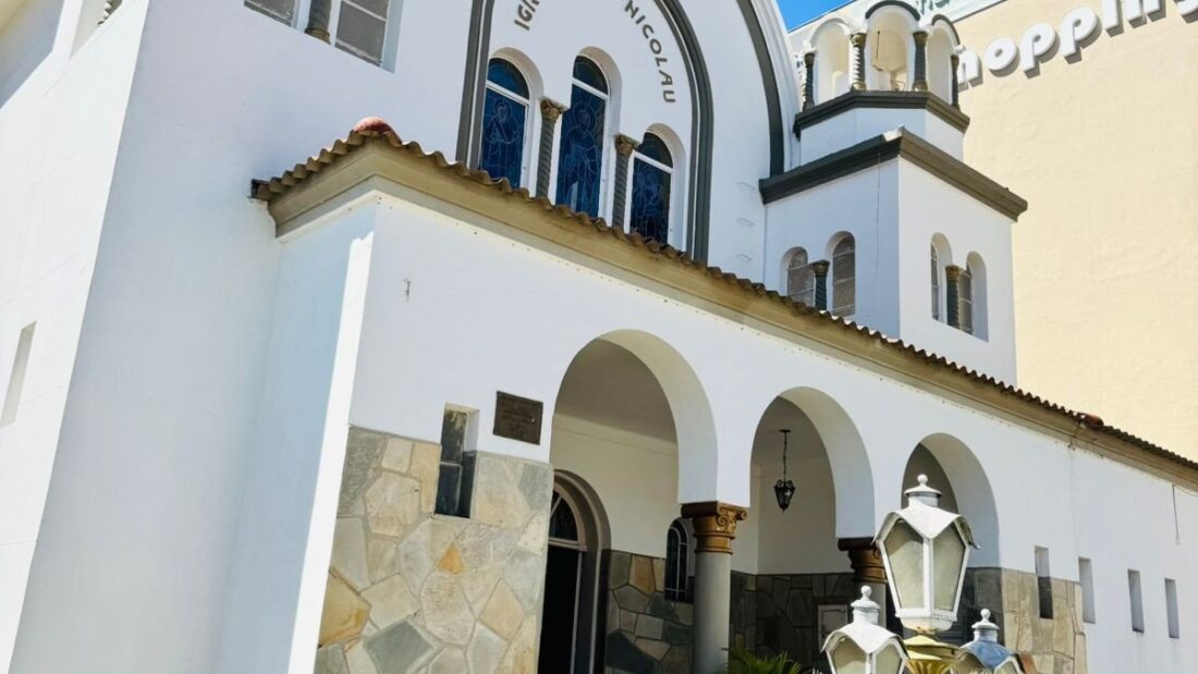 Fachada branca da Igreja Ortodoxa São Nicolau, em Goiânia, com arcos, cúpulas pequenas e cruzes no alto, e uma planta em primeiro plano
