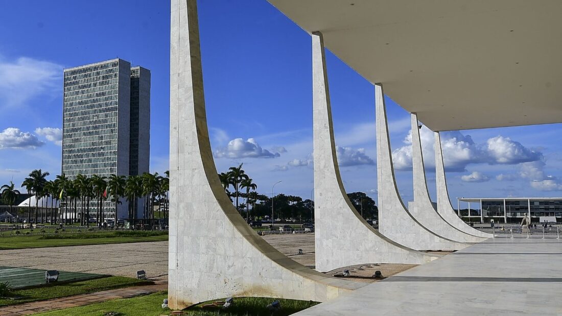 Colunas brancas curvas da fachada do Supremo Tribunal Federal, em Brasília, com um prédio alto ao fundo e céu azul com nuvens