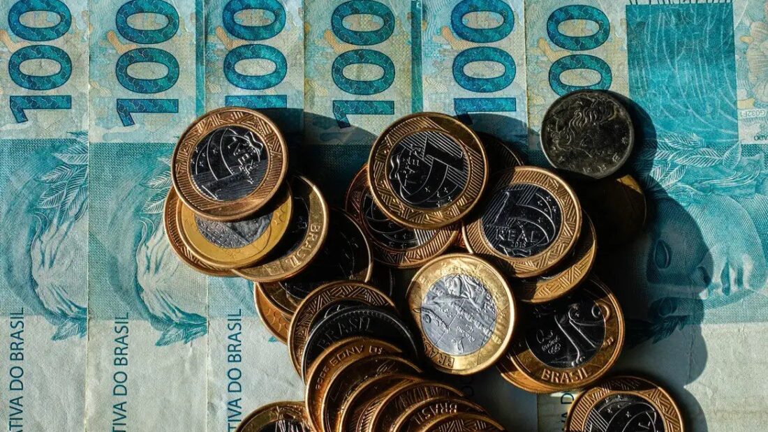 Moedas de um real e de centavos empilhadas sobre cédulas de cem reais