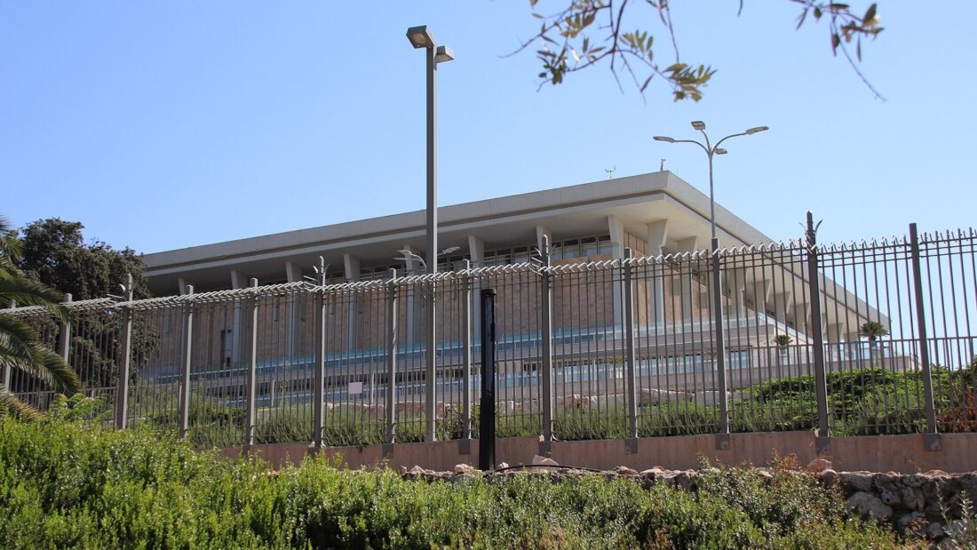 Prédio retangular do Knesset, o Parlamento de Israel, em Jerusalém, visto atrás de uma grade, com vegetação e postes em primeiro plano