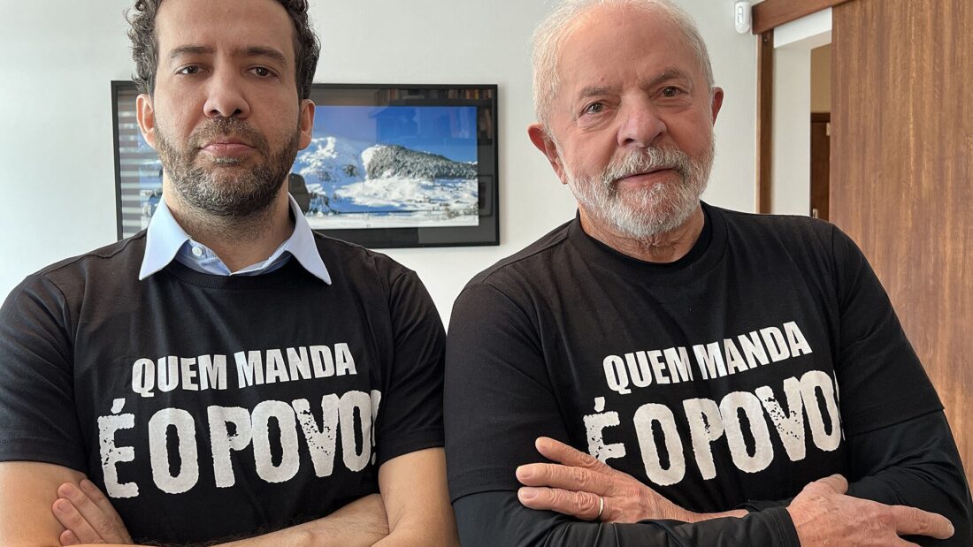 André Janones e Lula lado a lado, de braços cruzados, com camisetas pretas com a frase “Quem manda é o povo!”