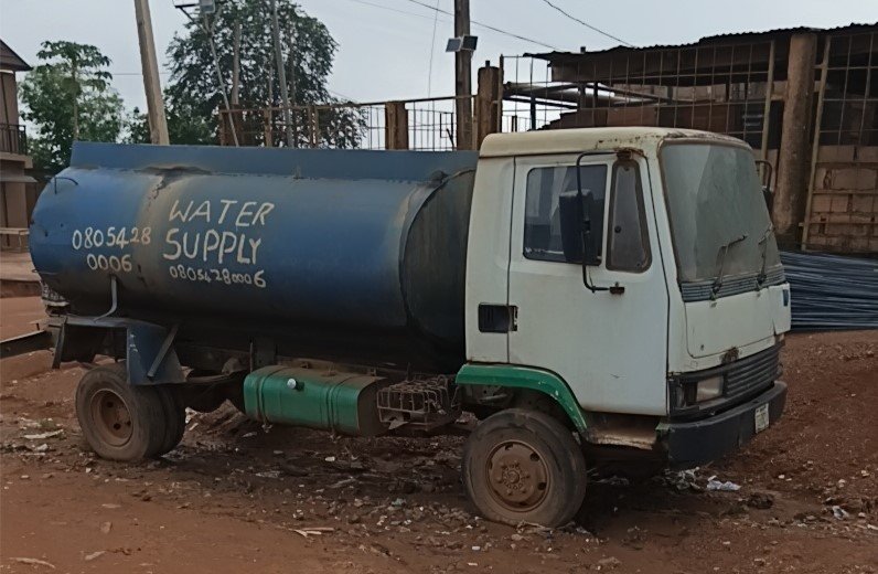 Caminhão-pipa azul e branco, com a inscrição Water Supply no tanque, estacionado em rua de terra