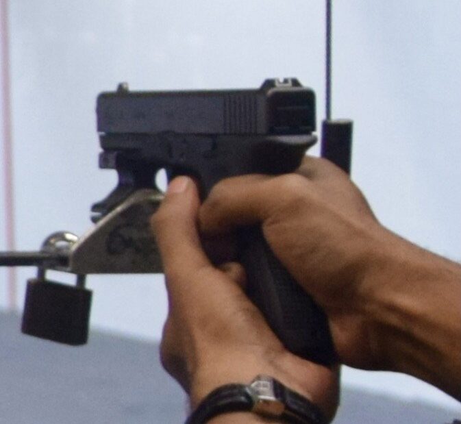 Mão segura uma pistola Glock preta, apontada para a frente, em um estande de tiro
