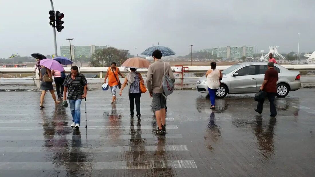 Pedestres com guarda-chuvas atravessam uma faixa de pedestres em avenida molhada durante chuva em Brasília