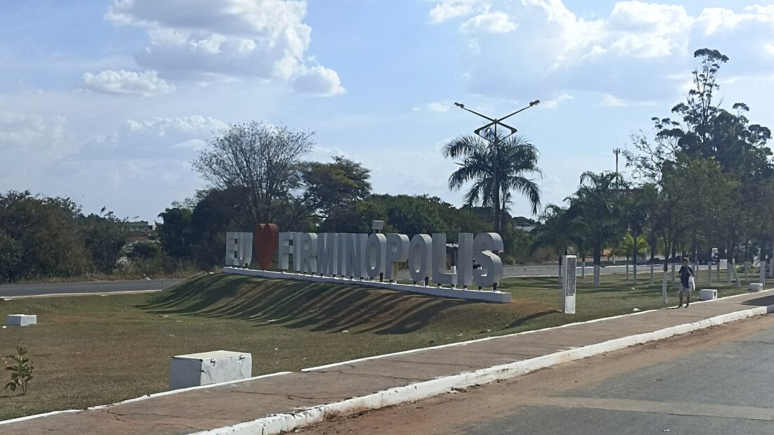 Letreiro com o nome de Firminópolis em um canteiro gramado na entrada da cidade, com árvores ao fundo