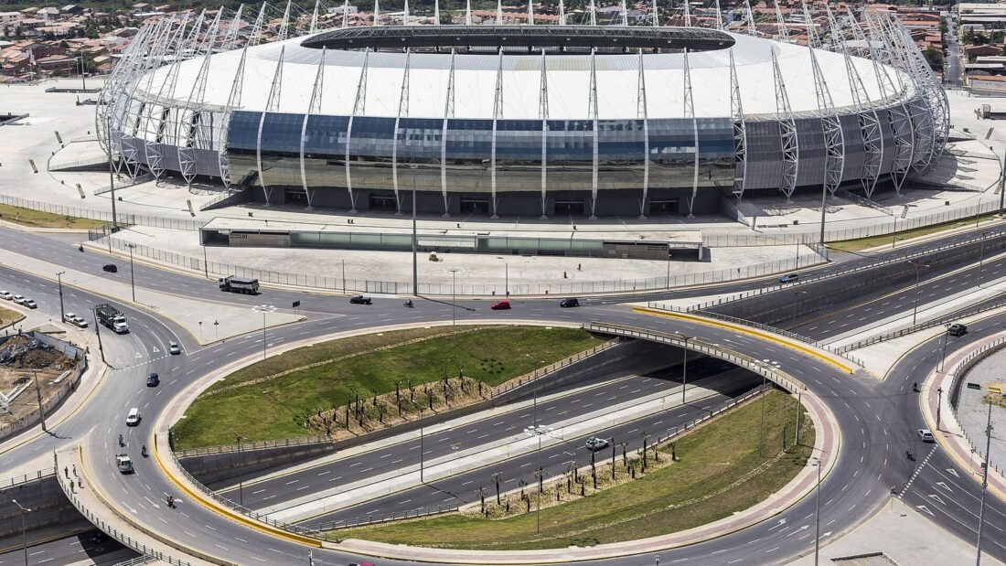 Vista aérea da Arena Castelão, em Fortaleza, com a cobertura metálica do estádio e um viaduto com avenidas e rotatória em primeiro plano