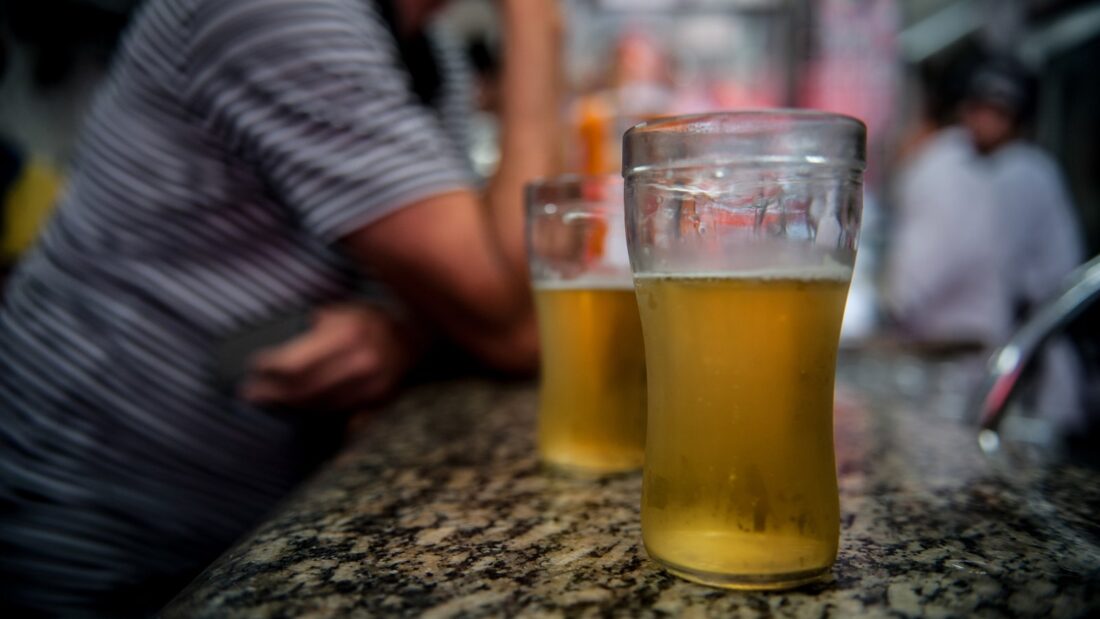 Dois copos de cerveja sobre o balcão de granito de um bar, com clientes desfocados ao fundo