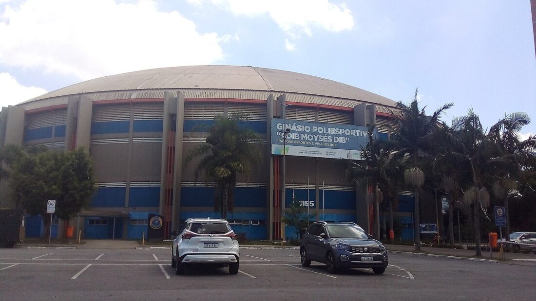 Fachada de um grande ginásio poliesportivo de cobertura arredondada, com carros estacionados à frente e palmeiras