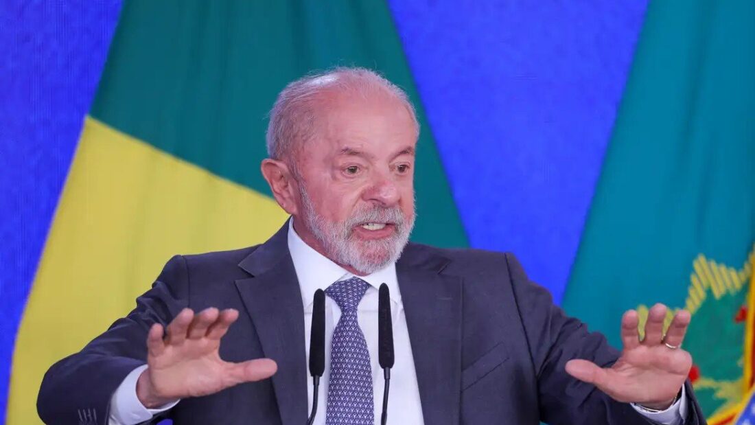 O presidente Lula, de terno escuro e gravata, gesticula com as duas mãos abertas ao discursar em frente a bandeiras do Brasil no Palácio do Planalto