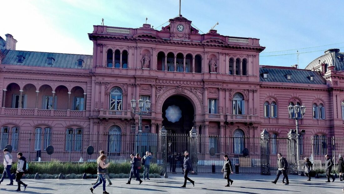 Fachada rosada da Casa Rosada, sede do governo argentino, com pedestres caminhando na praça em frente sob céu azul