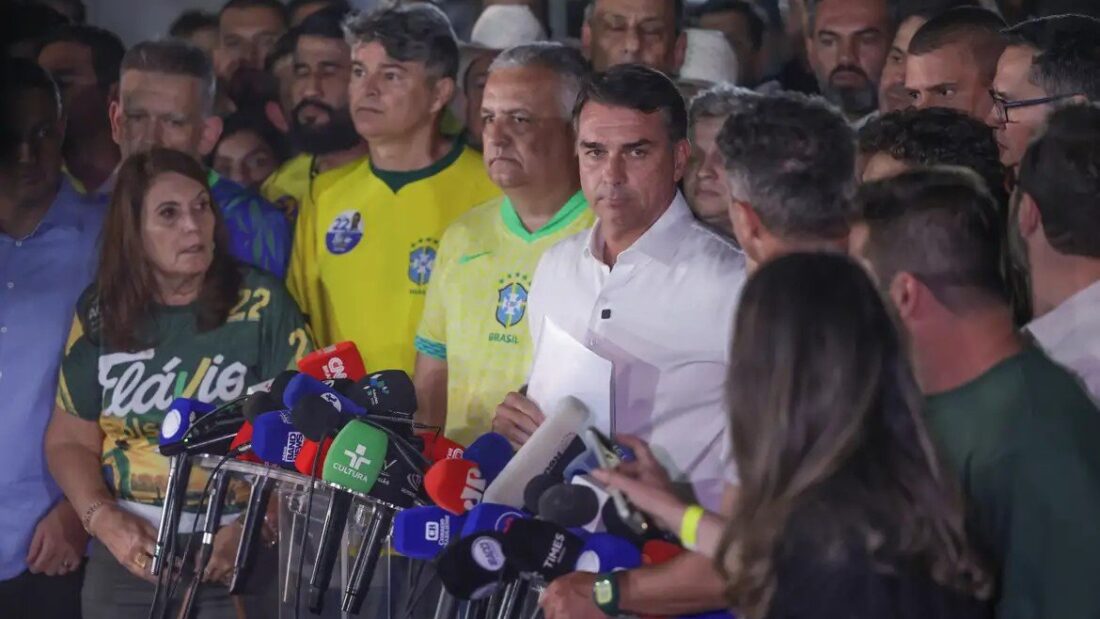 O senador Flávio Bolsonaro, de camisa branca, fala a jornalistas cercado de apoiadores com camisas da seleção e microfones de emissoras