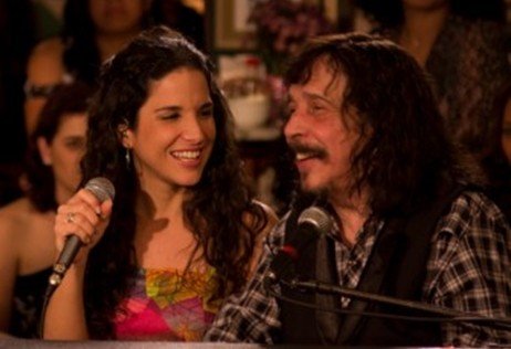 Benito di Paula, de cabelos longos e bigode, sorrindo ao lado da cantora Verônica Ferriani, que canta ao microfone, num ambiente de luz baixa e tons quentes