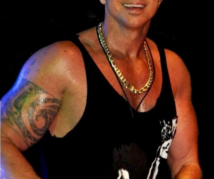 O cantor Netinho sorri no palco, de boné e camiseta regata preta