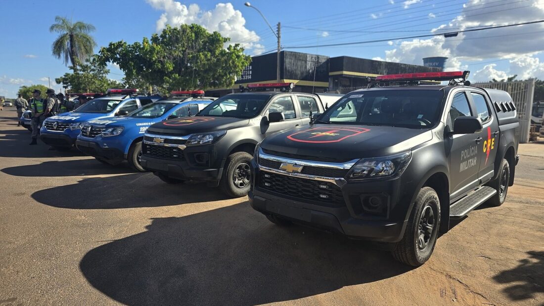 Caminhonetes pretas e azuis da Polícia Militar de Goiás estacionadas em fila em um pátio de terra