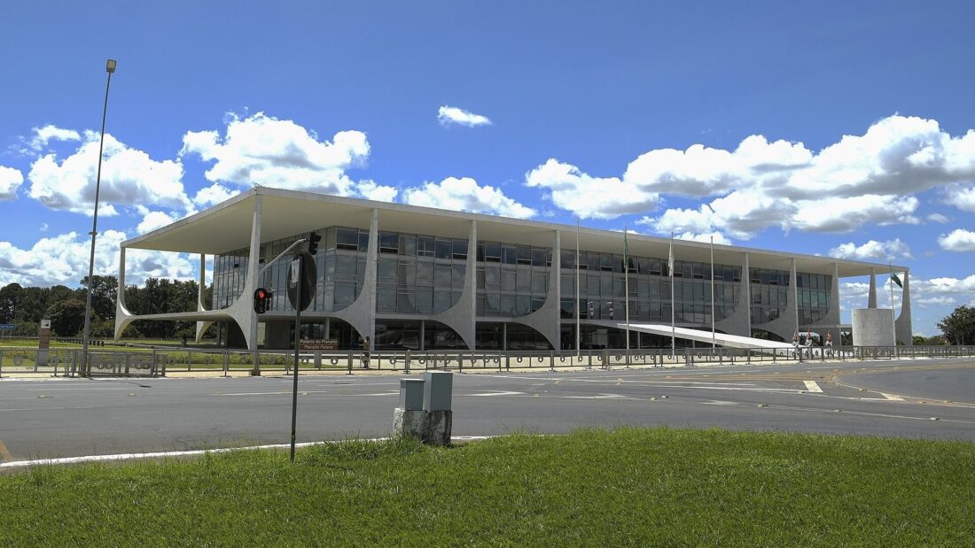 Fachada do Palácio do Planalto, em Brasília, prédio baixo e envidraçado com colunas brancas curvas, visto de uma pista vazia sob céu azul com nuvens