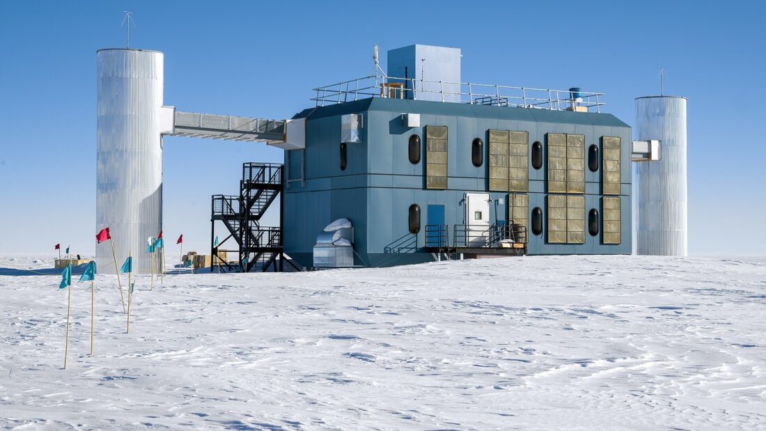 Prédio azul elevado sobre pilares, ladeado por duas torres cilíndricas metálicas, em meio à planície de neve do Polo Sul