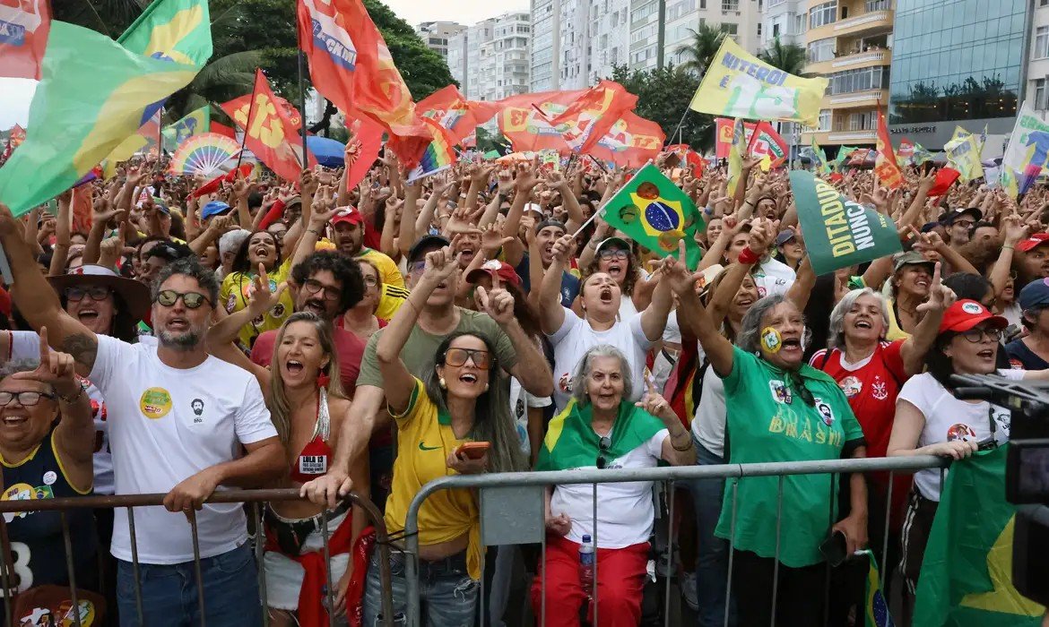 Multidão com bandeiras vermelhas e do Brasil, muitas pessoas com camisetas de campanha, erguendo os braços e gritando diante de uma grade na orla de Copacabana