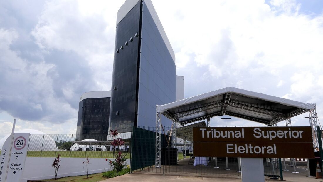 Prédio do Tribunal Superior Eleitoral, em Brasília, com torre de vidro escuro ao fundo e placa com o nome do tribunal sob uma cobertura na entrada