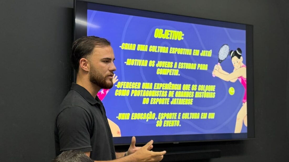 Homem de camisa preta apresenta, em um telão, os objetivos das Olimpíadas Escolares de Jataí durante reunião