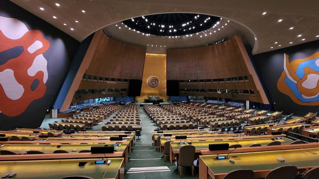 Salão da Assembleia Geral da ONU, em Nova York, visto do alto, com fileiras de mesas e cadeiras vazias, o painel dourado com o emblema das Nações Unidas ao fundo e grandes murais coloridos nas paredes laterais