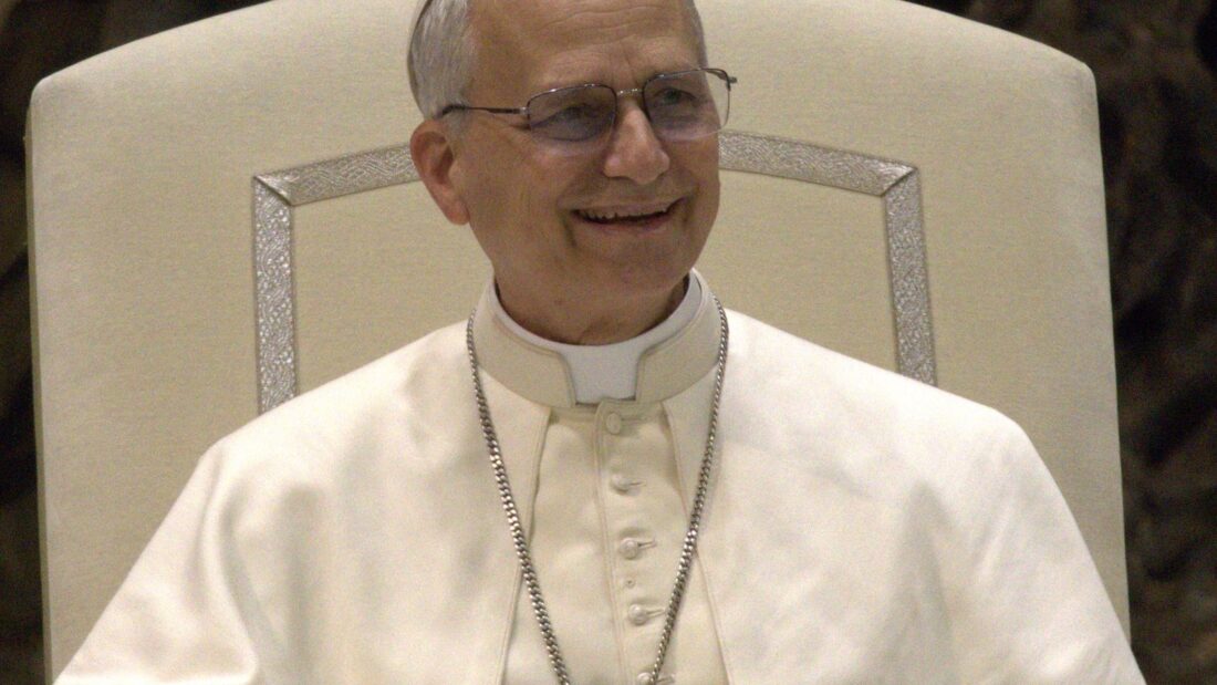 O papa Leão XIV, de batina branca, solidéu e crucifixo no peito, sorrindo sentado em uma poltrona clara