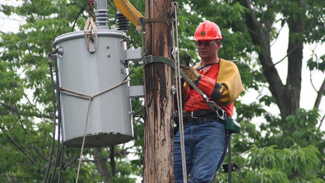 Eletricista com capacete e equipamento de segurança trabalha no alto de um poste, trocando um transformador de energia