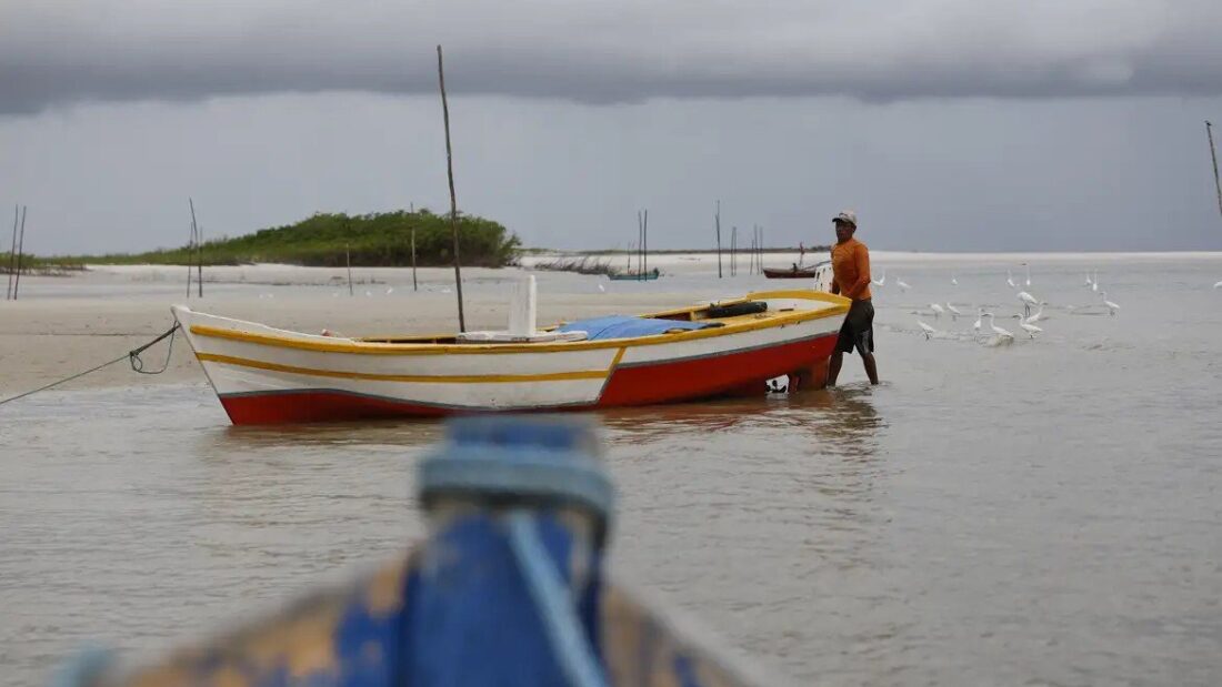 Pescador de pé na água rasa ao lado de um barco de madeira vermelho e branco, com aves e vegetação ao fundo