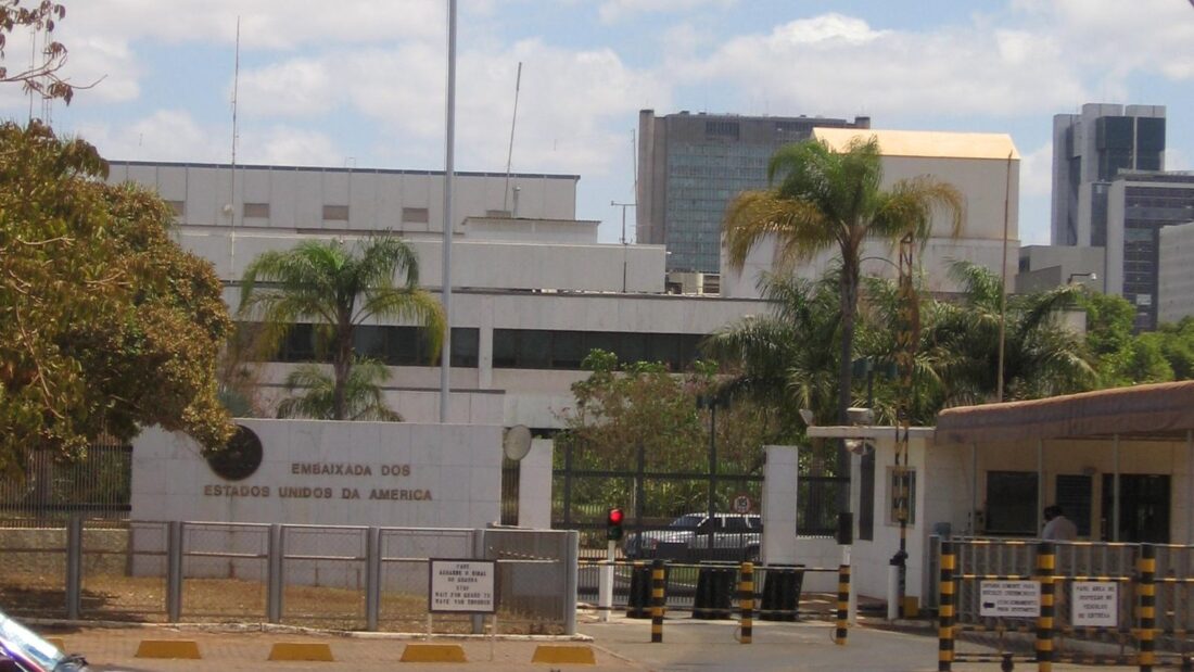 Entrada da Embaixada dos Estados Unidos em Brasília, com guarita, cones de sinalização, muro baixo e a bandeira norte-americana hasteada