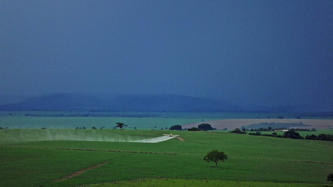 Avião agrícola faz aplicação sobre lavoura verde de soja sob céu carregado, com serra ao fundo e árvore isolada no campo